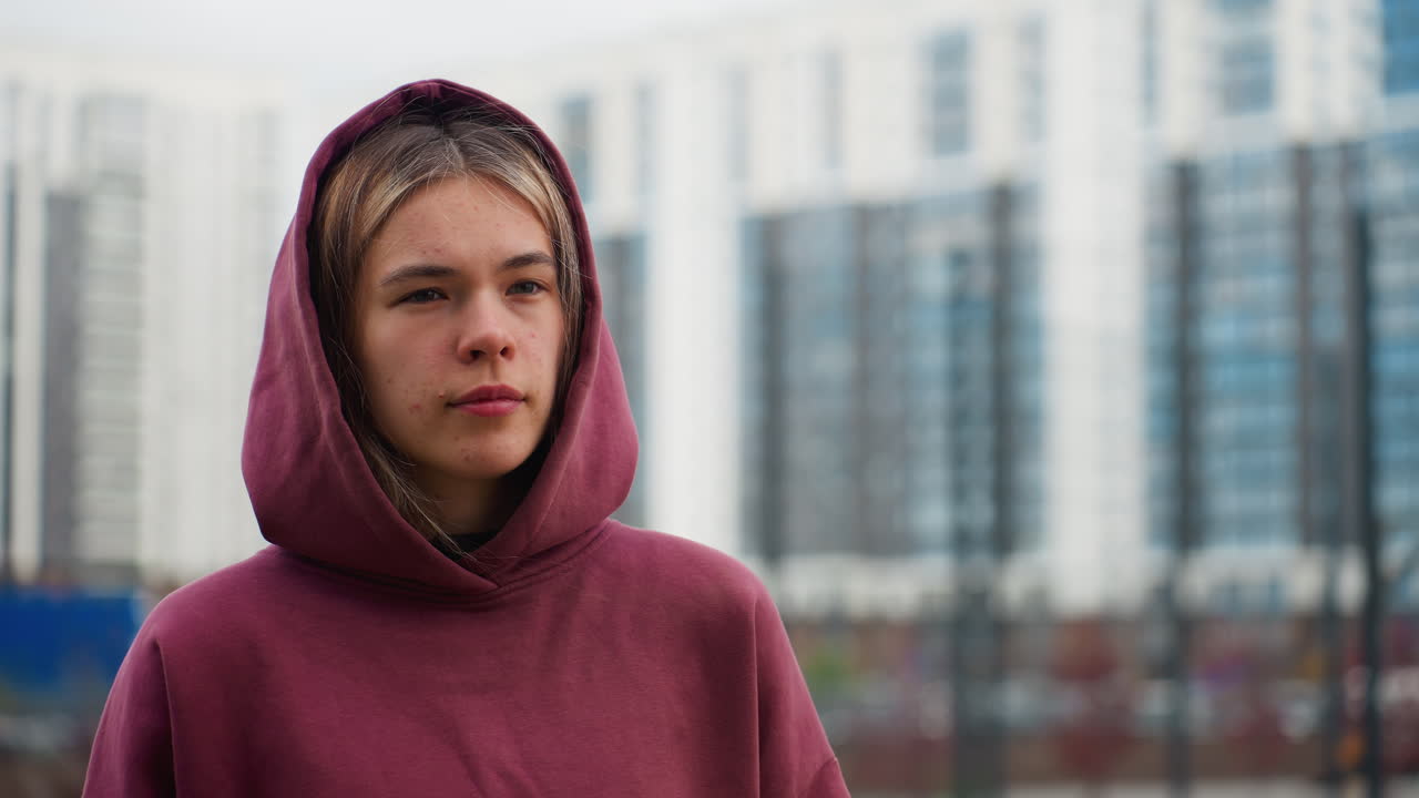 Youthful woman strolling urban walkway while glancing sideways past modern high rise buildings and spring flowers lining pathway in outdoor city environment under overcast sky feeling calm