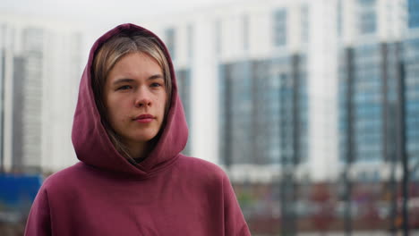 youthful woman strolling urban walkway while glancing sideways past modern high rise buildings and spring flowers lining pathway in outdoor city environment under overcast sky feeling calm
