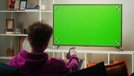 man watching television in living room. young guy sitting on sofa against tv with chroma green screen, back view. male person switching channels using remote controller.