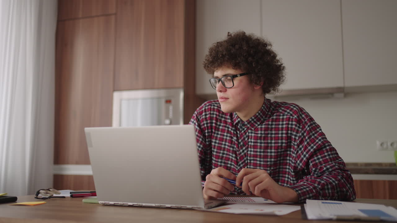 Curly haired Male student attractive young boy in glasses is studying at home using laptop typing writing in notebook. College student using laptop computer watching distance online learning seminar