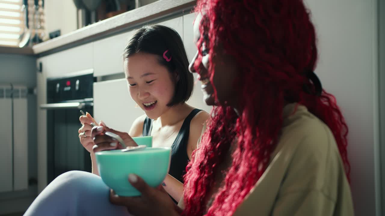 Two beautiful diverse friends eat corn flakes from a bowl sitting on the floor in the kitchen and laugh