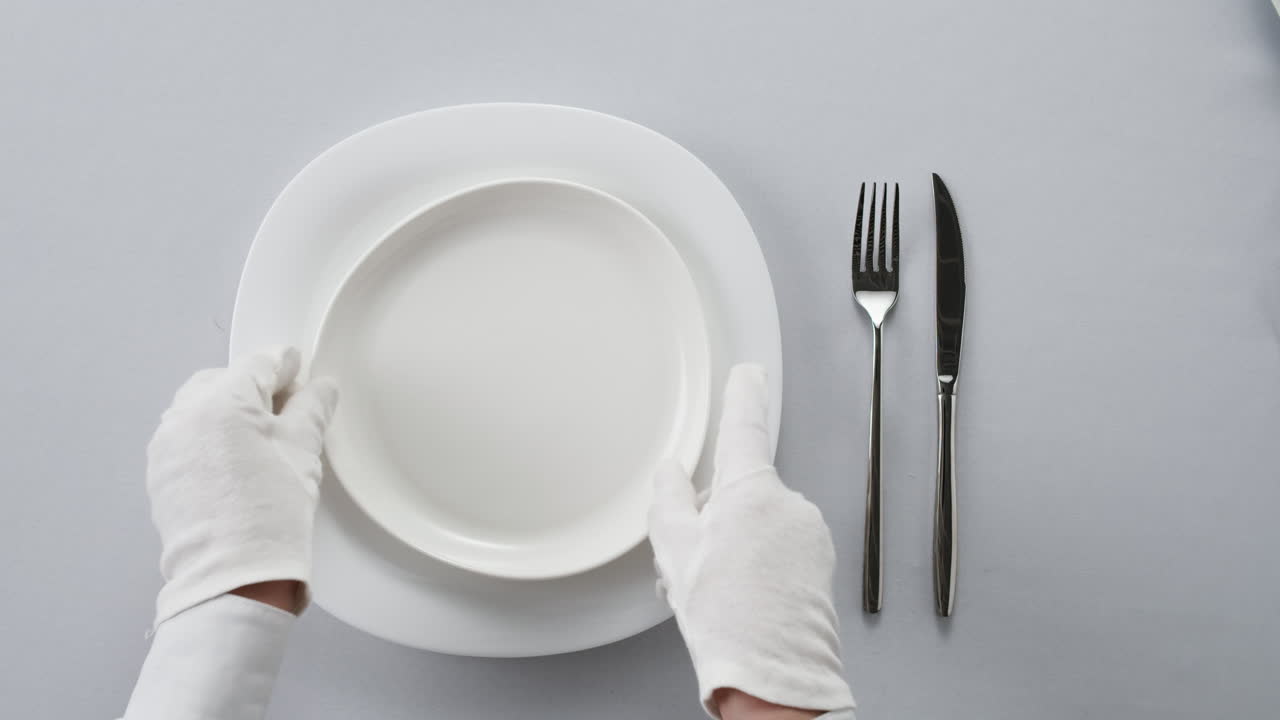 Professional table setting with white gloves. Plates, fork, knife, and glass arranged on a white table.