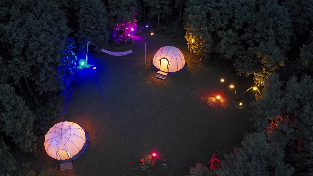 Aerial view of a campsite with glowing geodesic domes, illuminated by colorful lights at night, creating a magical and enchanting atmosphere in the forest