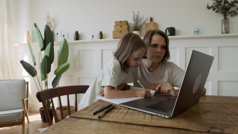 a pretty blonde little girl does her homework with the help of her mother while searching for information online on the laptop