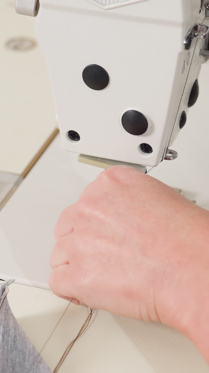 Primer plano de una máquina de coser que cose tela vaquera, manos que guían la aguja con un ritmo constante, prensatelas industrial que muerde un tejido grueso, bobina y carrete visibles, luz de estudio neutra, calma y concentración
