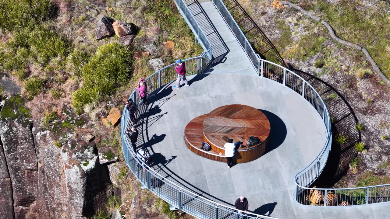 High-angle drone shot of people walking along a modern clifftop viewing platform in bright daylight, surrounded by rugged cliffs and natural bushland