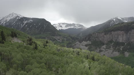 Forest-area-in-the-Rocky-Mountains-with-snowed-peaks-leading-to-a-canyon,-Colorado-USA,-Aerial-flyover-shot