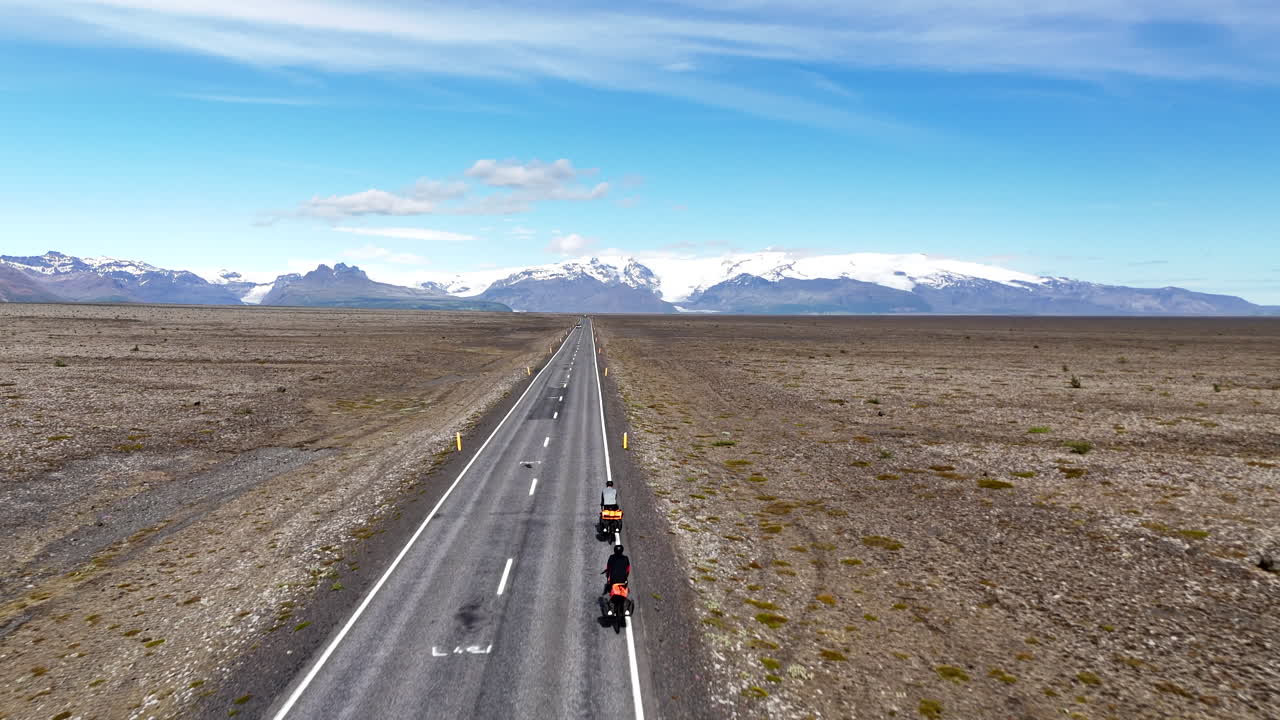Dronebillede i fugleperspektiv af to cyklister, der cykler på en øde vej nær Vatnajökull-gletsjeren i Island, og krydser en enorm vulkansk slette med sneklædte bjerge under en klar, blå himmel