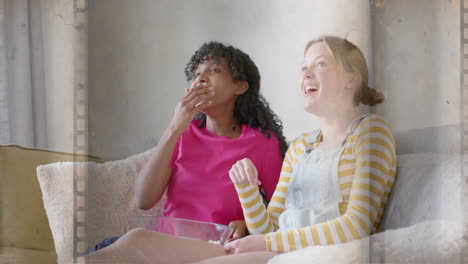 animation of film frame over two happy diverse teenage girls watching movie with popcorn at home