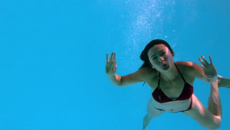 Happy-brunette-waving-at-camera-underwater-in-swimming-pool