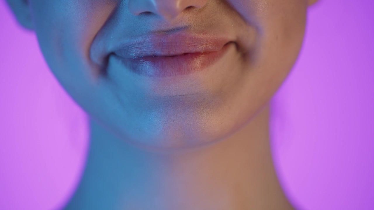 Macro ripresa 4K al rallentatore delle labbra sorridenti di una donna, sfondo viola vibrante con illuminazione soffusa blu e rosa sul suo viso, primo piano cinematografico e artistico, tonalità luminose e dettagli di colore vividi