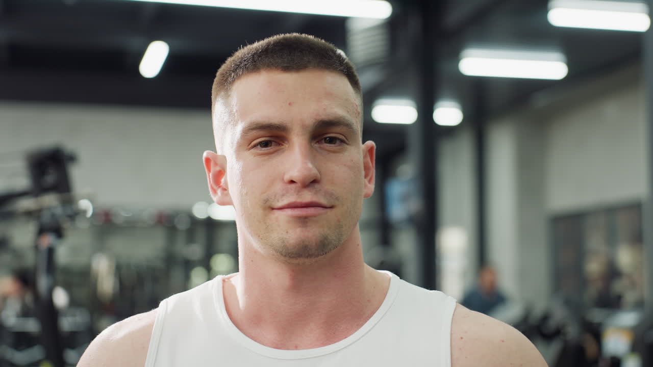Portrait view of gym instructor smiling inside modern gymnasium, wearing white tank top, standing between exercise machines while fluorescent ceiling lights create reflections, showing confidence