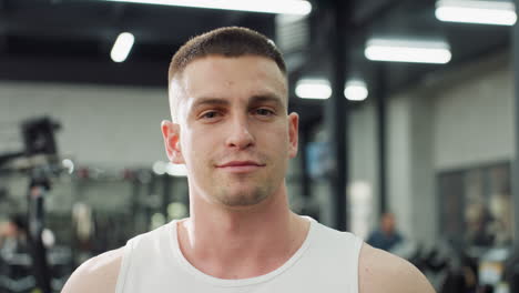 portrait view of gym instructor smiling inside modern gymnasium, wearing white tank top, standing between exercise machines while fluorescent ceiling lights create reflections, showing confidence