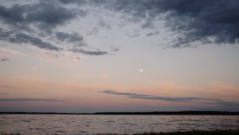 Ein-Zeitraffer-Des-Sonnenuntergangs-Mit-Blick-Auf-Einen-See,-Während-Sich-Wolken-Am-Himmel-Bewegen-Und-Im-Hintergrund-Der-Mond-Zu-Sehen-Ist