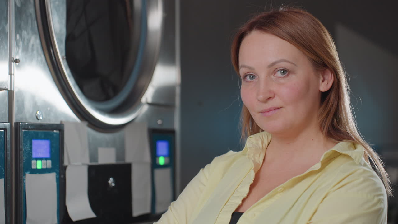 Service woman standing near industrial washer observing cycle, clothes visible inside drum, stainless control panels lit, staff monitoring laundry process in modern laundromat interior, side profile