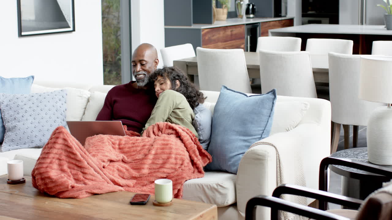 Happy biracial couple sitting on couch and using laptop at home, slow motion