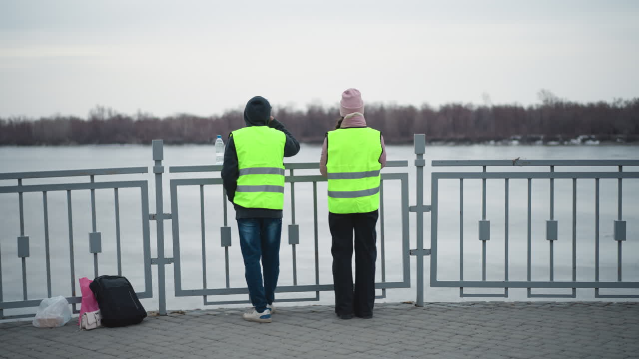 Two people wearing reflective safety vests stand by riverside railing in cold weather, looking across frozen water with backpacks and bags on ground, overcast sky, and bare trees in distance