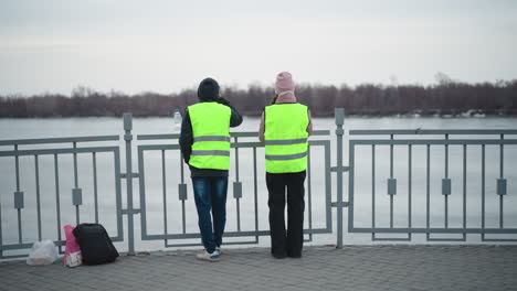 two people wearing reflective safety vests stand by riverside railing in cold weather, looking across frozen water with backpacks and bags on ground, overcast sky, and bare trees in distance