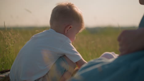 a back view of a little boy on a scarf in a grassy field, leaning forward as he plays. a blurred background features a blue flowing gown and a partially visible boy lying nearby