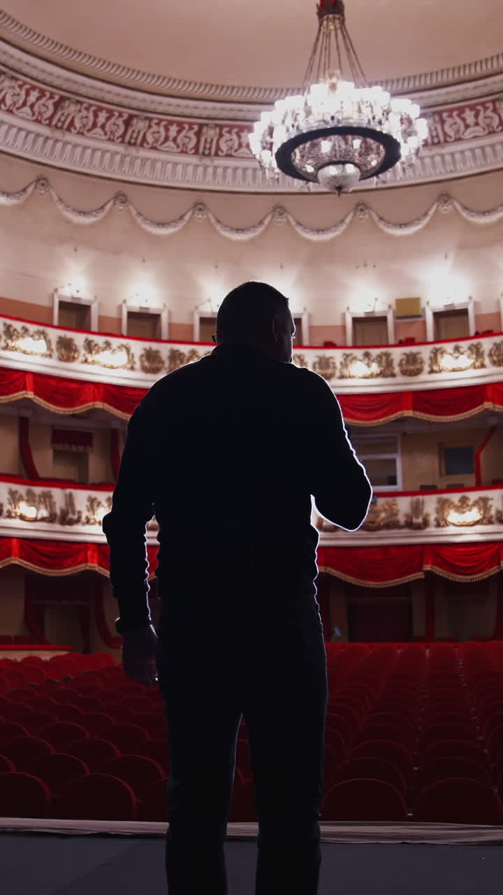 Actor on stage in spotlight. Active man standing on stage of a classical theatre with rows of red chairs without audience. Back view. Vertical video
