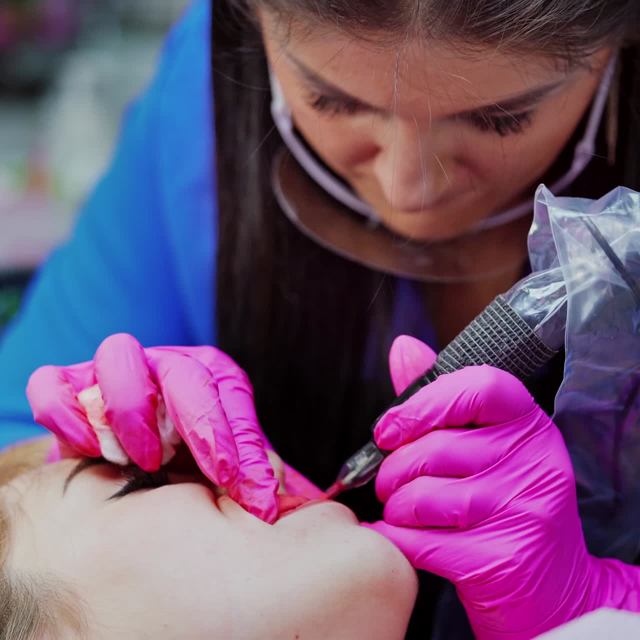 Lips tattoo. Cosmetologist in protective gloves applying permanent make-up on lips. Professional tattooist working with special tattooer machine.