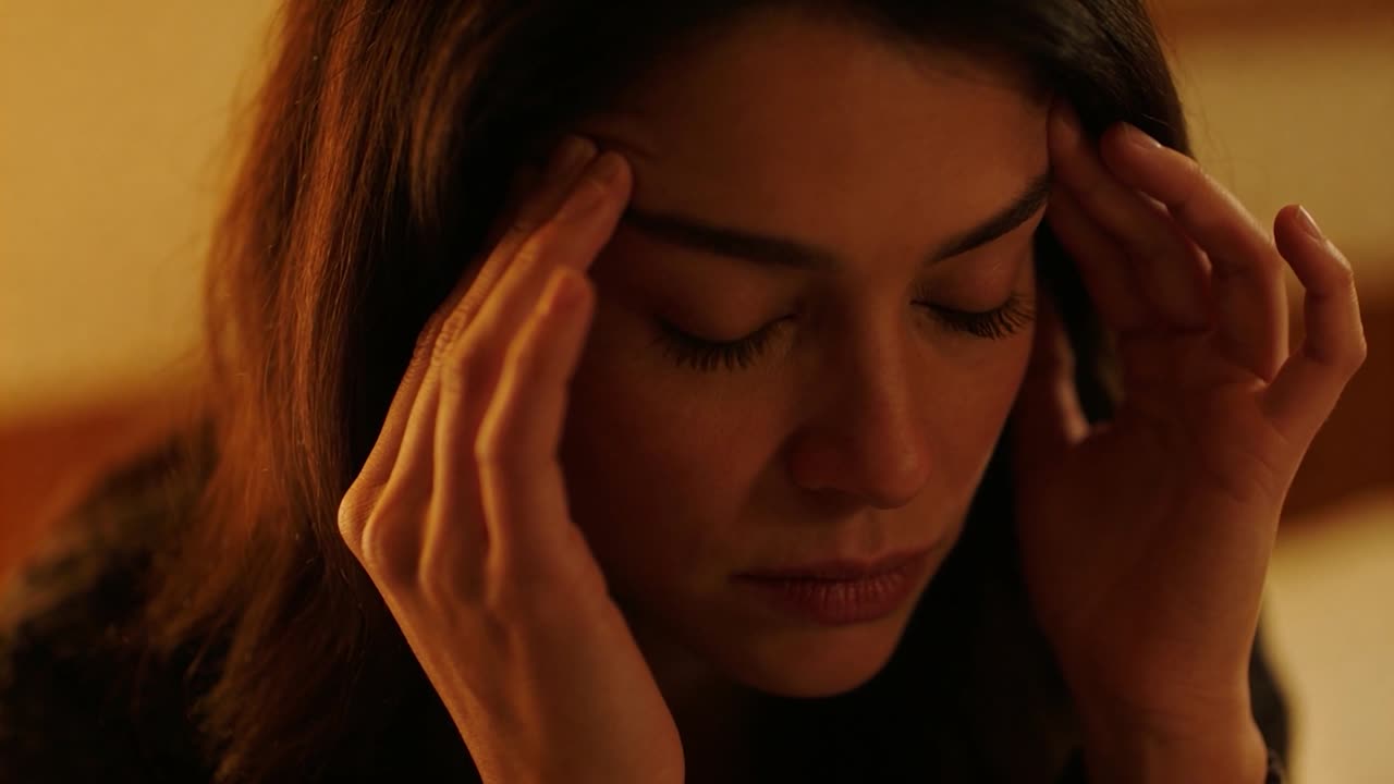 Donna con lunghi capelli scuri che preme i polpastrelli sulle tempie, cercando sollievo dal mal di testa in camera da letto.
