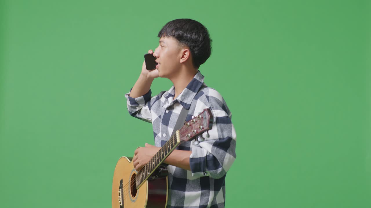 초록색 화면 배경에 서서 기타를 들고 스마트폰으로 대화하는 아시아 십대의 측면 시각