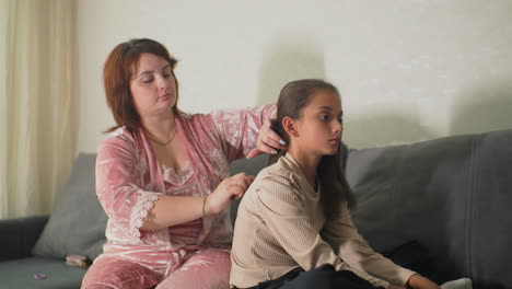 Mother brushing daughter’s hair at home on couch in cozy living room with natural light shining through curtains. Tender, intimate family moment showing care, connection, and love between them