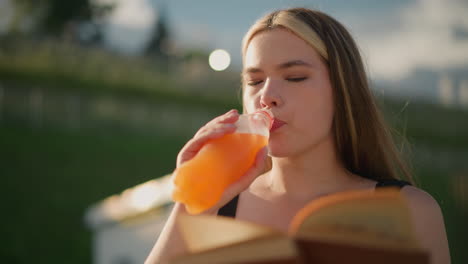 mujer sentada al aire libre, sosteniendo un libro en una mano mientras beba jugo de una botella en la otra, continúa leyendo, con el libro ligeramente borroso y un fondo sereno y borroso