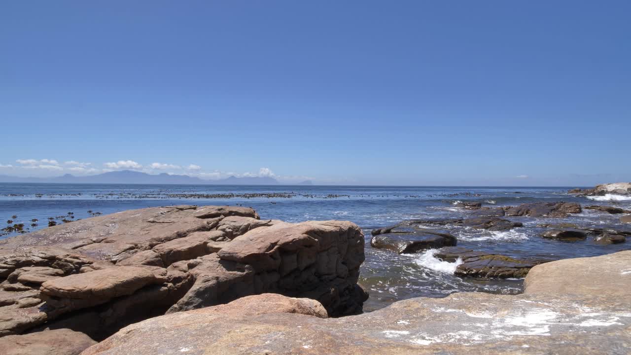 Weitläufiger Meerblick mit verwitterten Felsformationen und sanften Wellen, die das Ufer am Cape Point umspülen, mit Blick auf die False Bay und fernen Bergen am Horizont