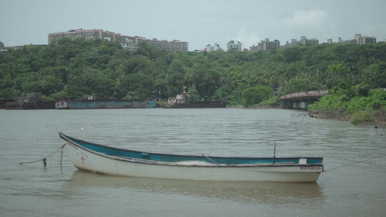 Tranquil static shot frames a wooden boat anchored on calm Chicalim Bay, with a backdrop of dense green hills, tropical trees, and distant buildings in Goa, India