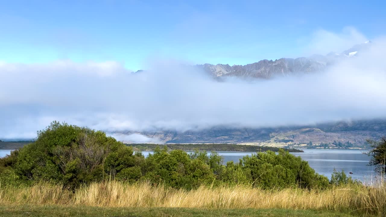 Clouds move across Lake Wakatipu, revealing scenic views of mountains and greenery under bright daylight in Queenstown, New Zealand