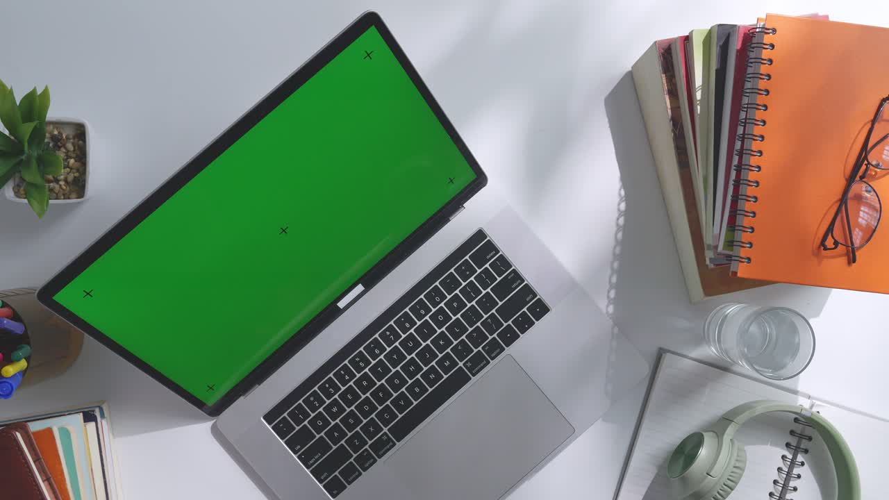 vista de arriba hacia abajo de una computadora portátil con pantalla verde simulada pantalla chromakey en un escritorio de oficina de madera al lado de la libreta con bolígrafos, gafas y un vaso de agua.