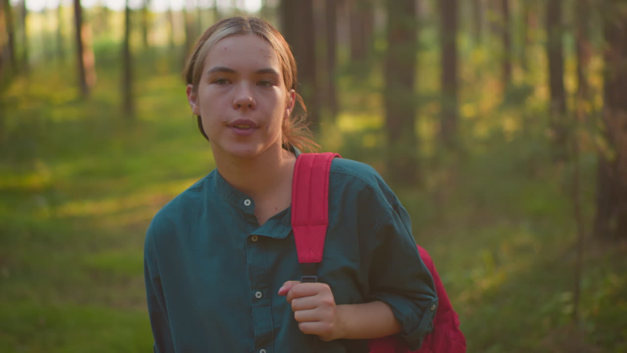 excursionista agotado caminando a través de un bosque pacífico, ajustando la correa de la bolsa roja en el hombro, mirando pensativo a su alrededor, bañado en la cálida luz del sol que arroja un suave resplandor en su cara