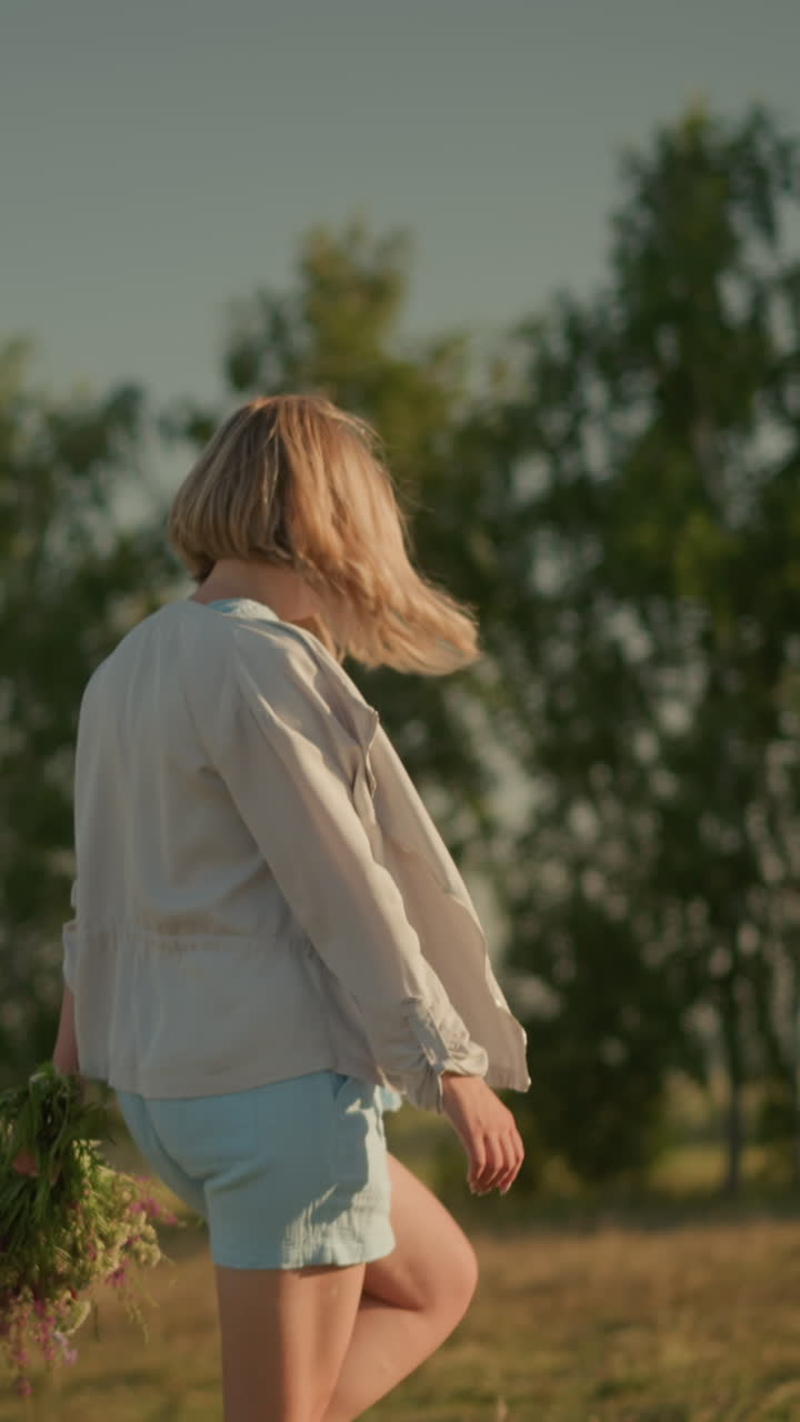 짧은 바지와 긴 팔을 가진 셔츠를 입은 여성이 은 날 손에 꽃을 들고 열린 을 걸어가고, 배경에는 흐릿한 나무와 초록색이 있습니다.