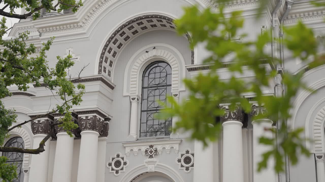 Vista detallada de la fachada de la iglesia de San Miguel Arcángel en Kaunas, con una arquitectura compleja y una exuberante vegetación. Captura el ambiente sereno en un día despejado.