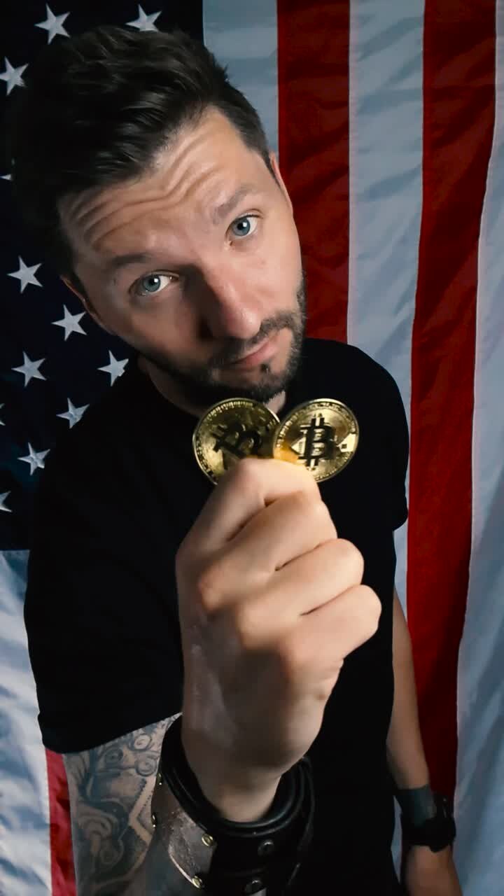 Tattooed man with a beard holding up and showing two golden and shiny Bitcoin cryprocurrency money coin to the camera while standing in front of vertical hanging American United states flag. Bullish.