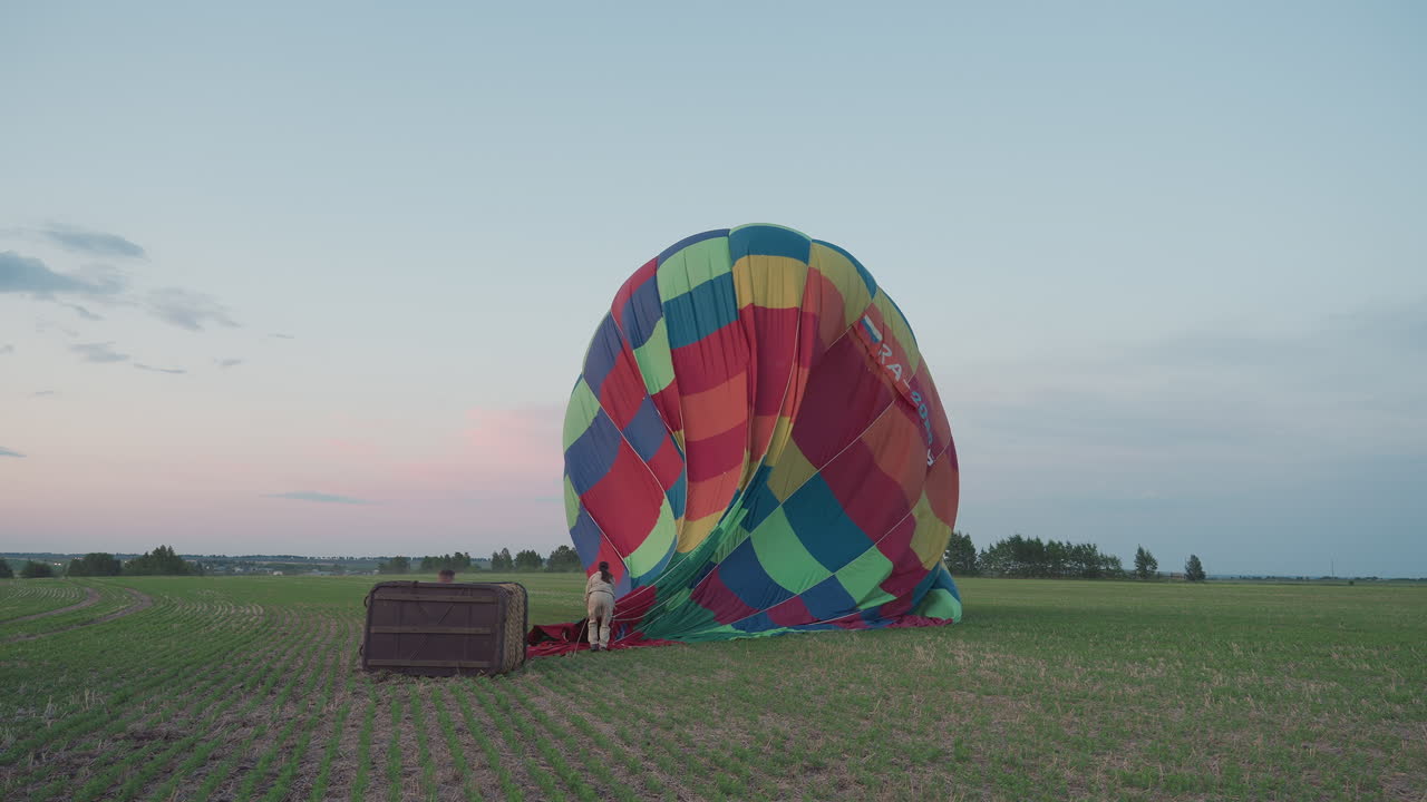 woman pulling deflated colorful hot air balloon canopy across green farmland toward waiting trailer under pastel dusk sky capturing teamwork and careful motion