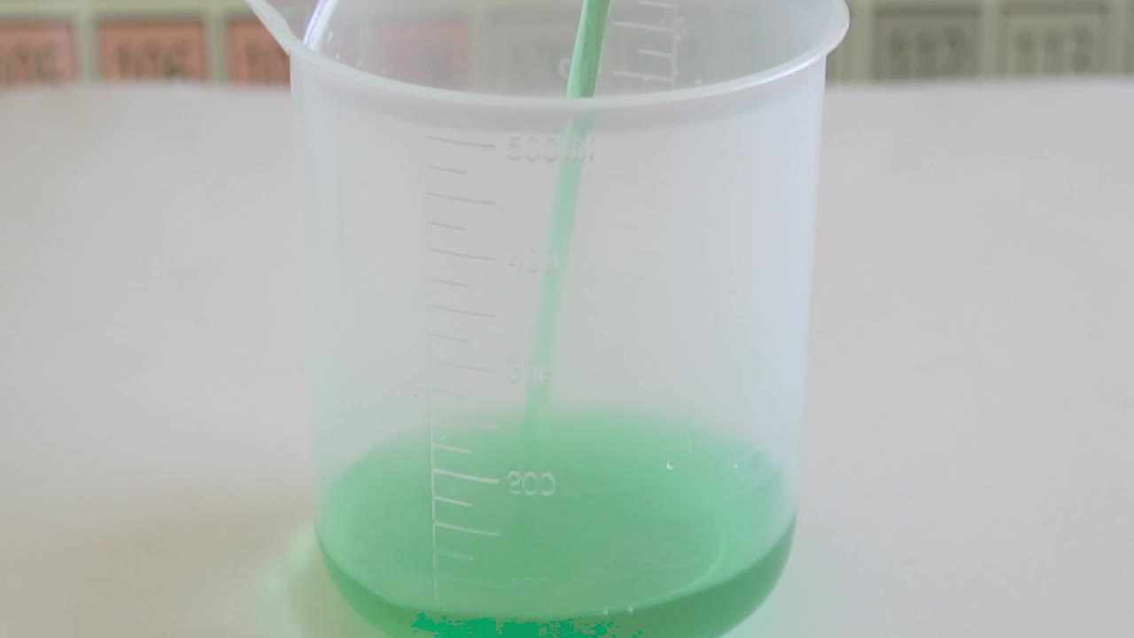 Chemicals are being poured into a beaker with periodic table in background .