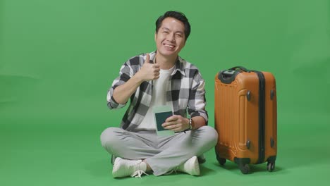 full body of asian male traveler with luggage and passport smiling and showing thumbs up gesture to the camera while sitting in the green screen background studio