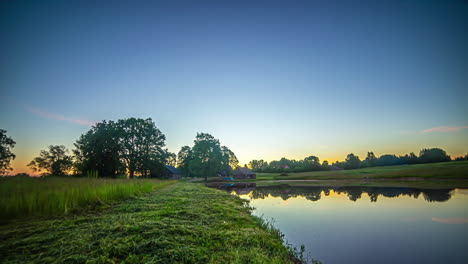 Tiro-De-Lapso-De-Tiempo-Del-Campo-Rural-Con-Vista-De-La-Cabaña-De-Madera-Al-Lado-De-Un-Lago-En-La-Distancia-Al-Amanecer