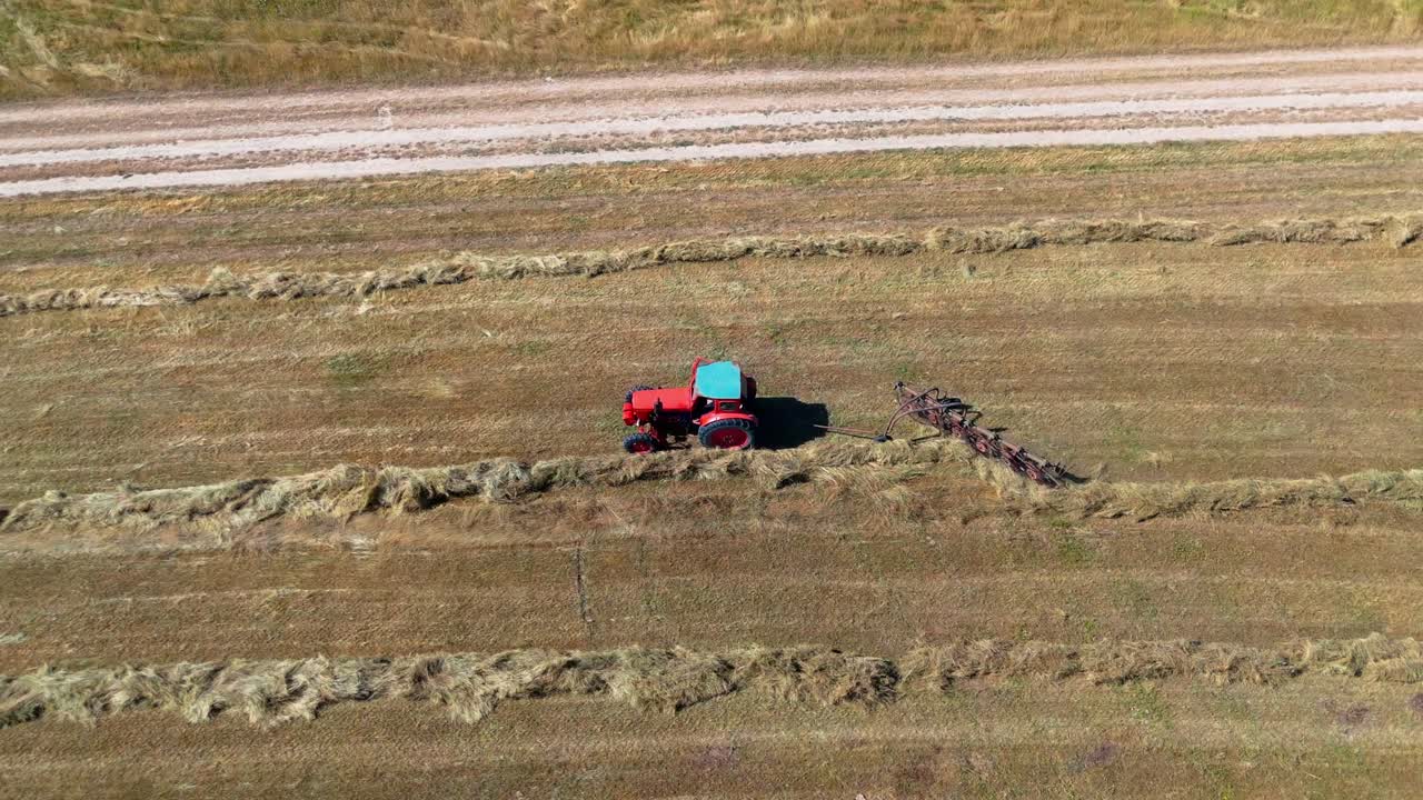 Top-down drone pan following a red tractor raking hay across cultivated farmland, emphasizing movement, symmetry, and agricultural patterns