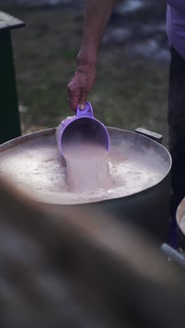 slow motion close up of hot chocolate milk being poured from a purple container into a large pot, with steam rising, suggesting an outdoor cooking scene for large number of persons