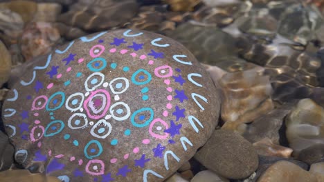 Close-up-shot-of-colored-old-stone-in-clear-water,-painted-by-children-in-nature---Kids-painting-and-distribute-it-on-the-forest-and-meadow-path