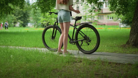 woman approaches black bicycle parked on paved path in lush green park, placing one glove on bike seat while wearing other glove, background features tall trees, and people walking in the distance