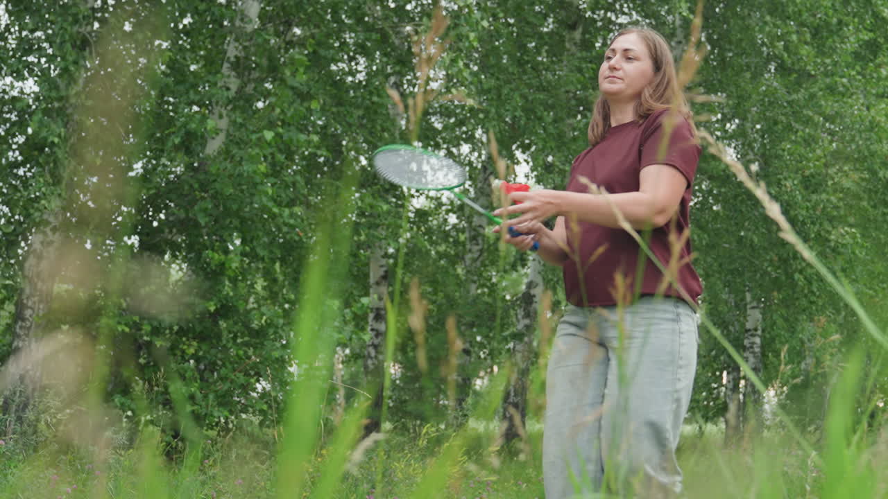 Una mujer blanca jugando al bádminton en un campo de hierba con hierba baja en primer plano; lanzamientos y capturas dinámicas con raqueta y volante; camiseta color granate y vaqueros que sugieren que la atleta está calentando; aficionada.