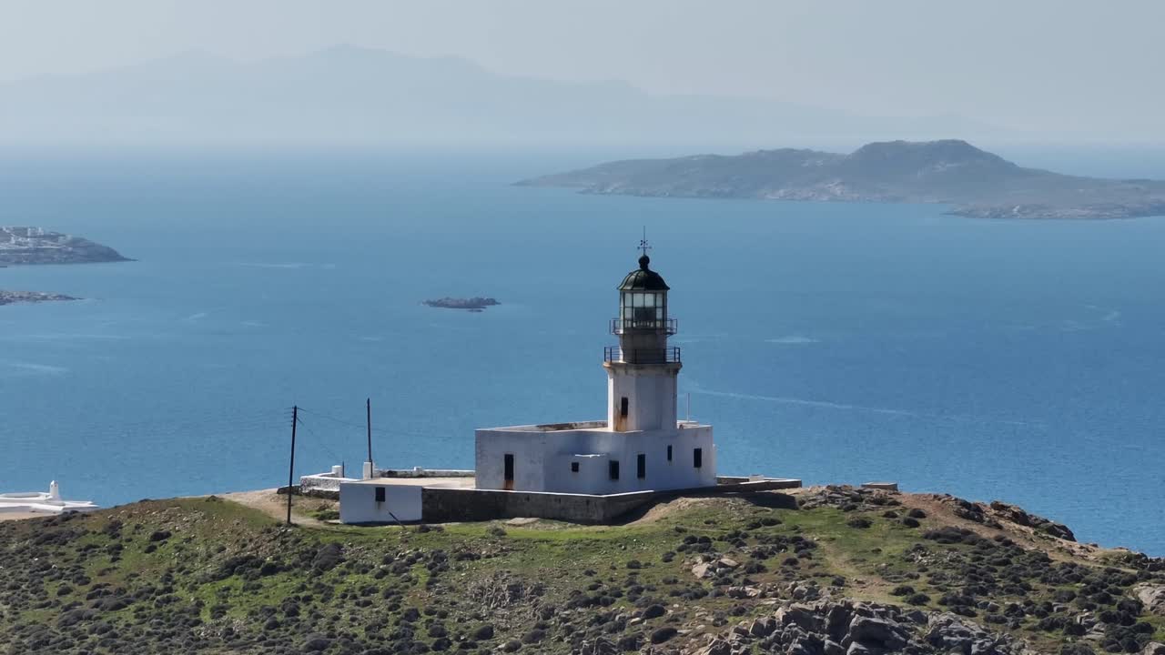 Drone orbit around Mykonos Lighthouse capturing stunning views of Delos island and Chora village under a bright sunny sky, showcasing the beauty of the Aegean Sea and Cyclades