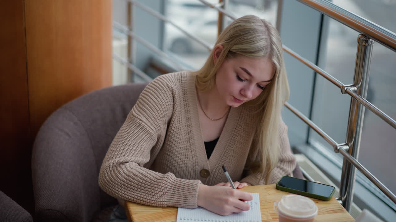 Lady writing in reflective mood while looking up. Background features winter scene with blurred view of parked cars and people moving about outside the window