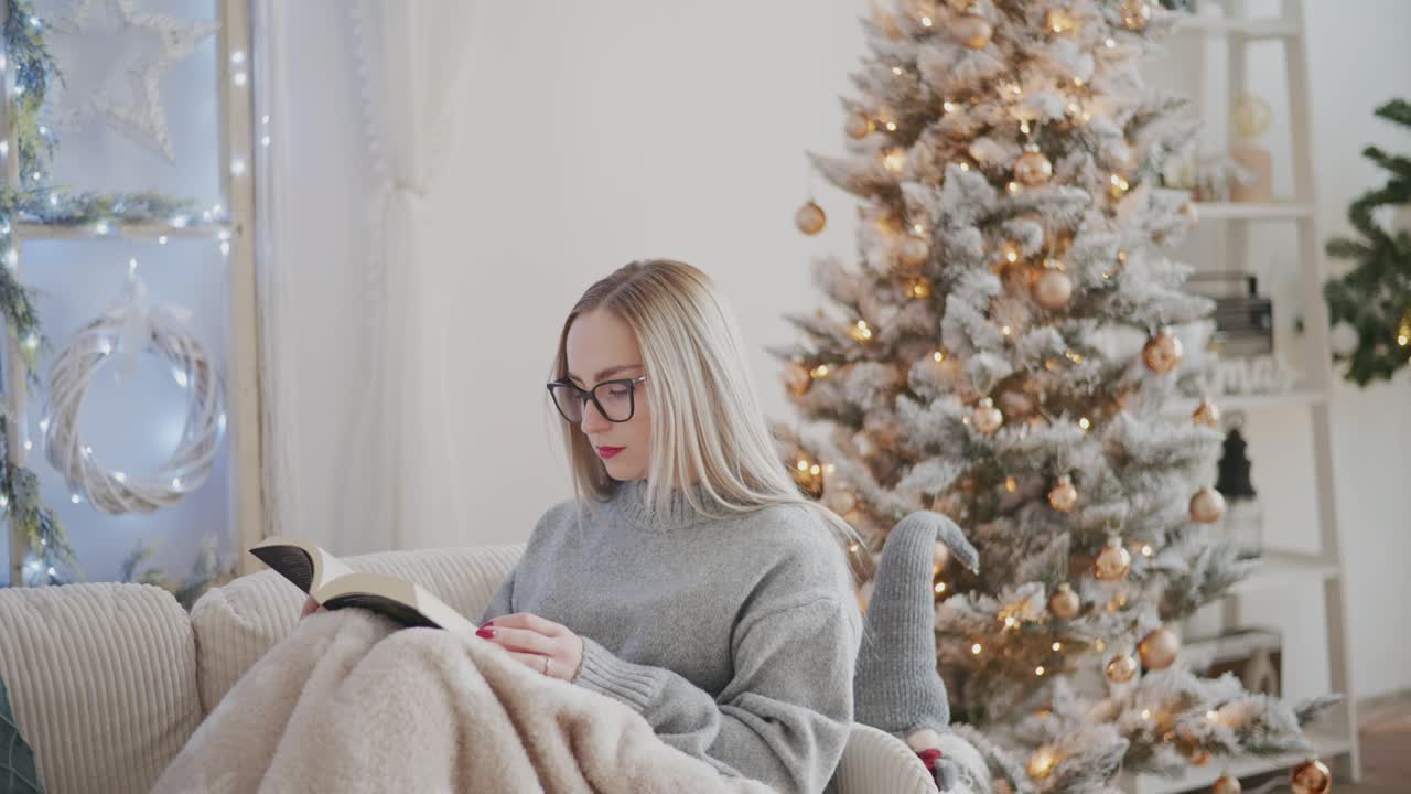 A woman in a cozy gray sweater reads a book while sitting on a sofa near a beautifully decorated Christmas tree. Warm fairy lights and festive ornaments create a peaceful holiday atmosphere at home