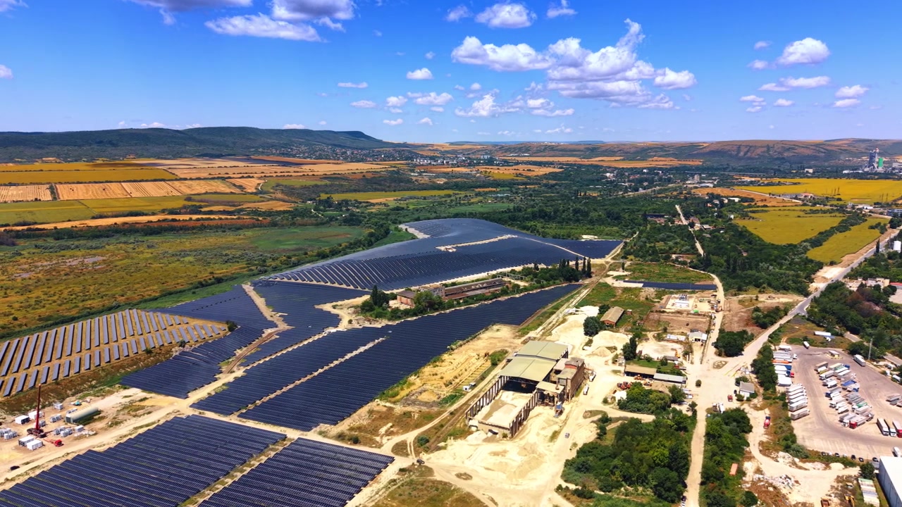 Vista aerea di un impianto fotovoltaico vicino a campi agricoli. L'inquadratura ampia mostra una schiera di pannelli solari, un paesaggio verde, camion parcheggiati nel piazzale, cielo blu con nuvole.